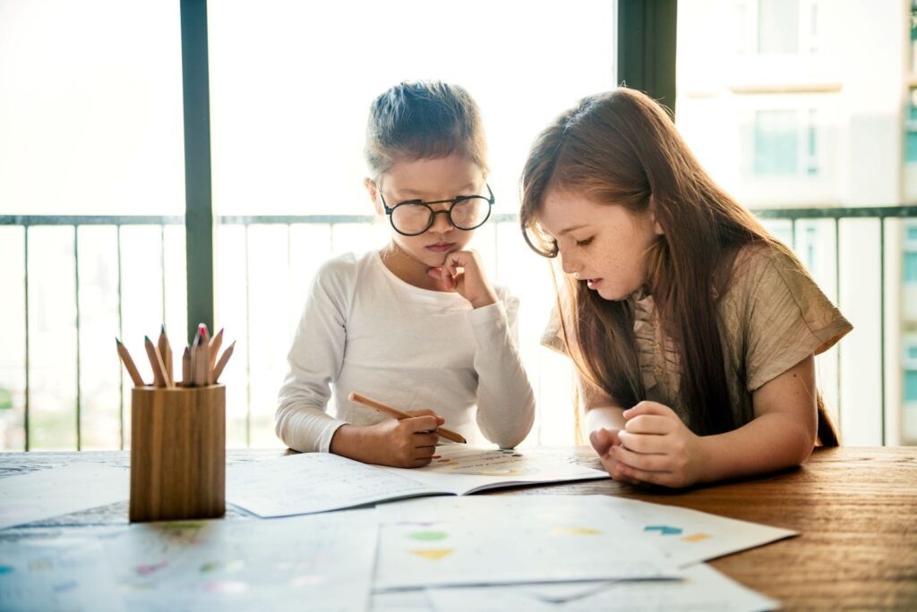 Two children studying together at a table with papers and pencils