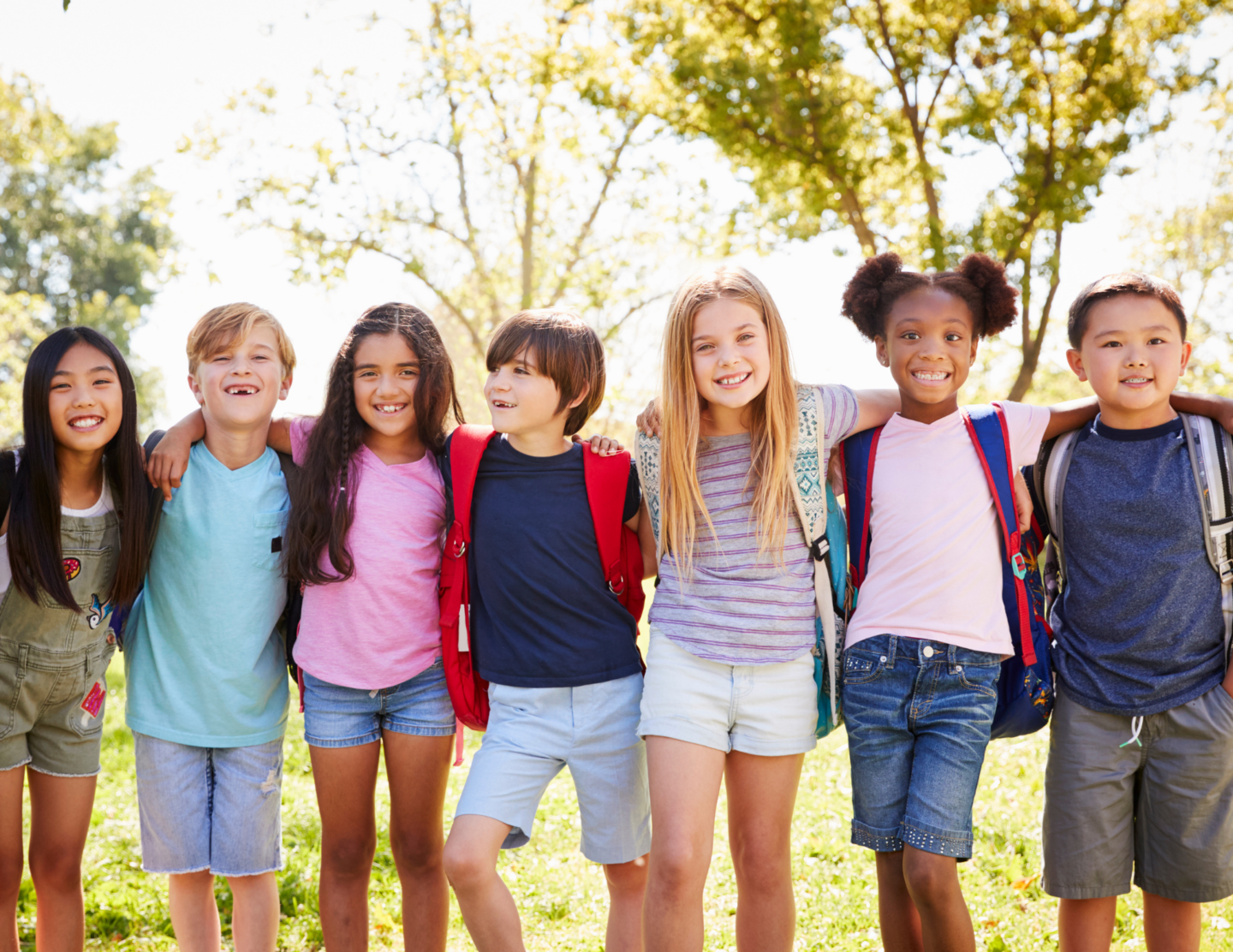 Group of smiling children with backpacks standing outdoors in a sunny park