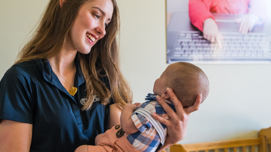 Smiling woman holding a baby in a nursery setting with a poster in the background