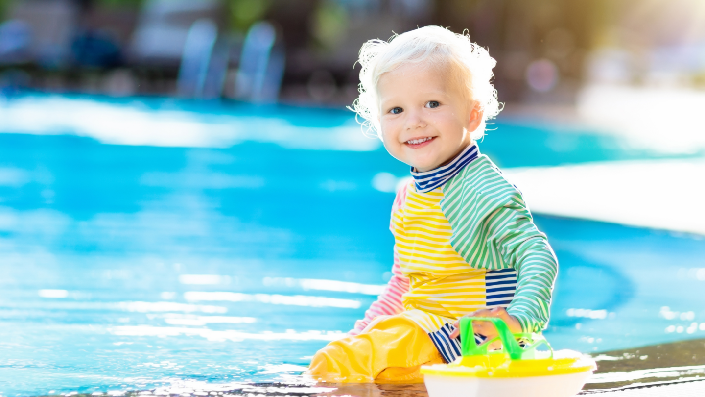 Toddler in colorful swimwear smiling by a pool with a toy boat