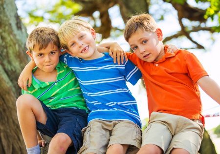 Three boys sitting outdoors on a tree branch smiling and wearing colorful shirts on a sunny day