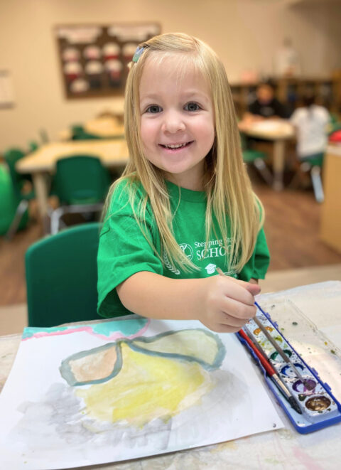 Young girl painting with watercolors in a classroom with green chairs.