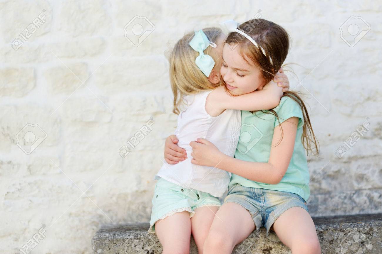 Two young girls hugging with joy on a stone bench against a light brick wall