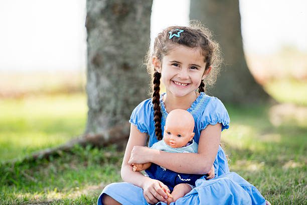 Young girl in blue dress holding a doll while sitting on grass outdoors