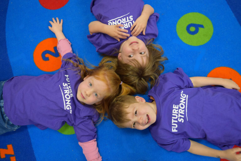 Three kids in purple Future Strong shirts lying on a colorful numbered rug smiling.