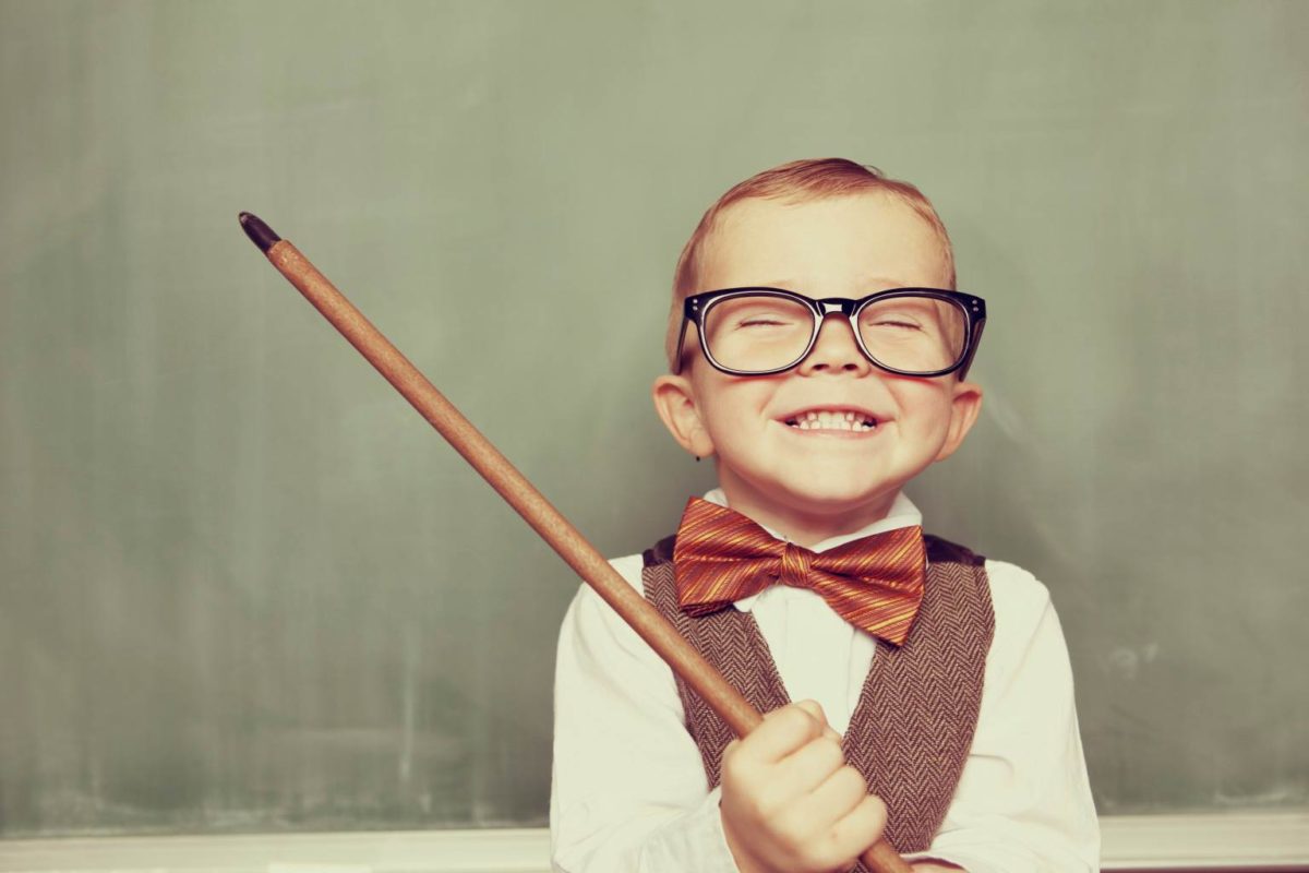 Smiling child with glasses and bow tie holding a pointer in front of a chalkboard