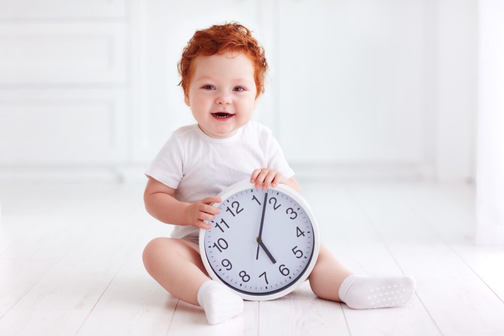 Smiling baby with red hair holds large clock on white floor background