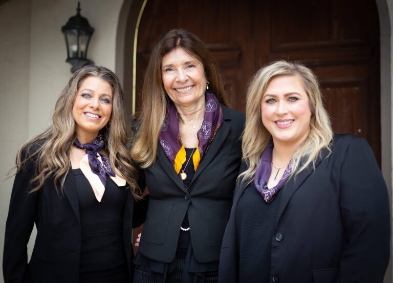 Three women in business attire smiling in front of a wooden door