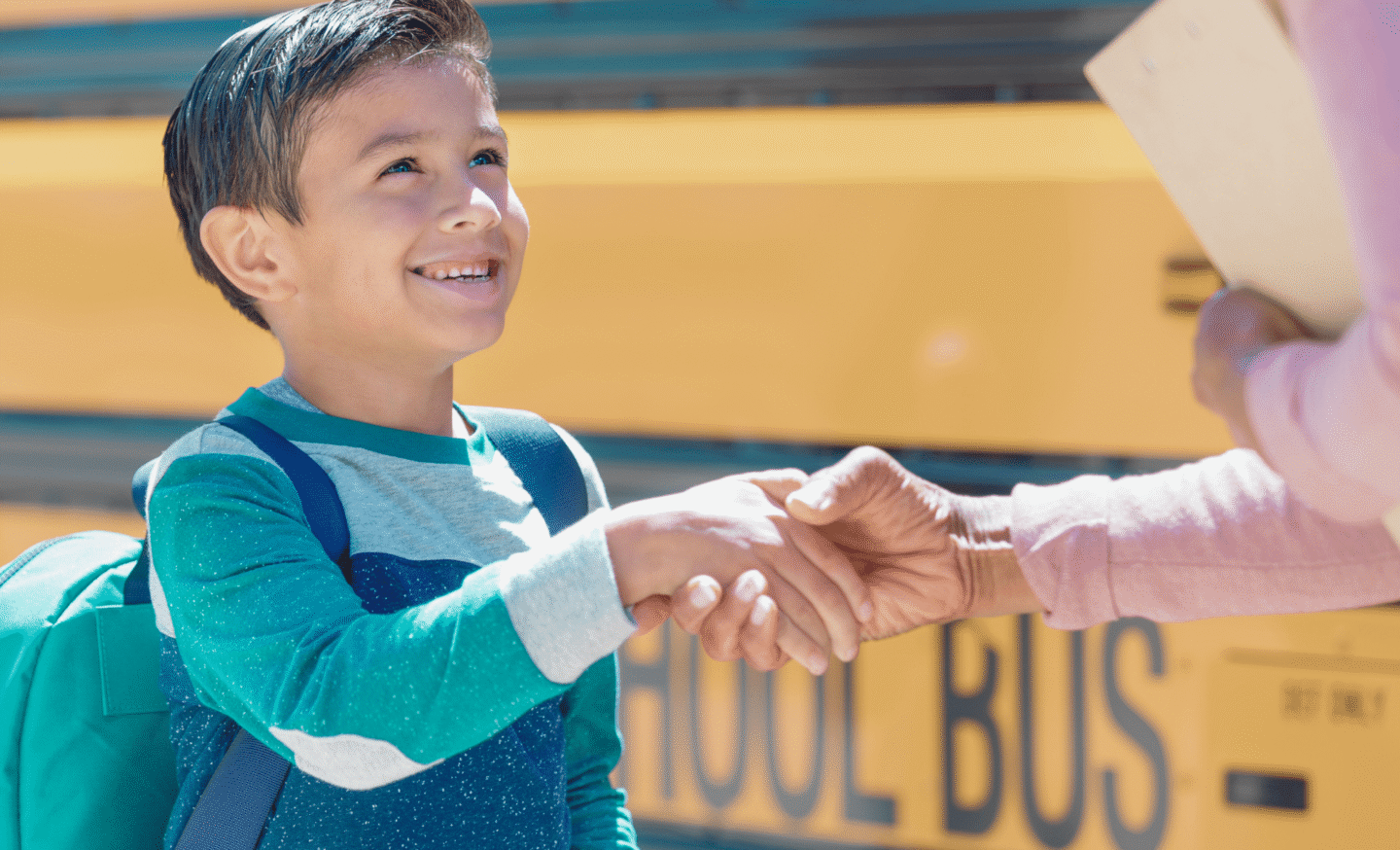 Boy with backpack smiling and shaking hands in front of a school bus