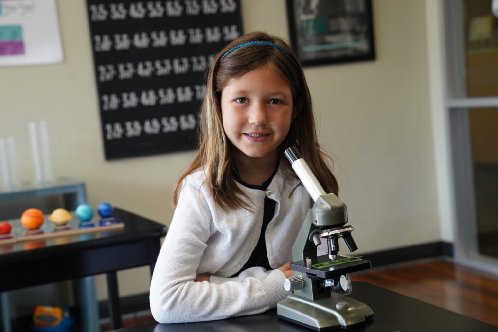 Young girl smiling and sitting at a table with a microscope in a science classroom