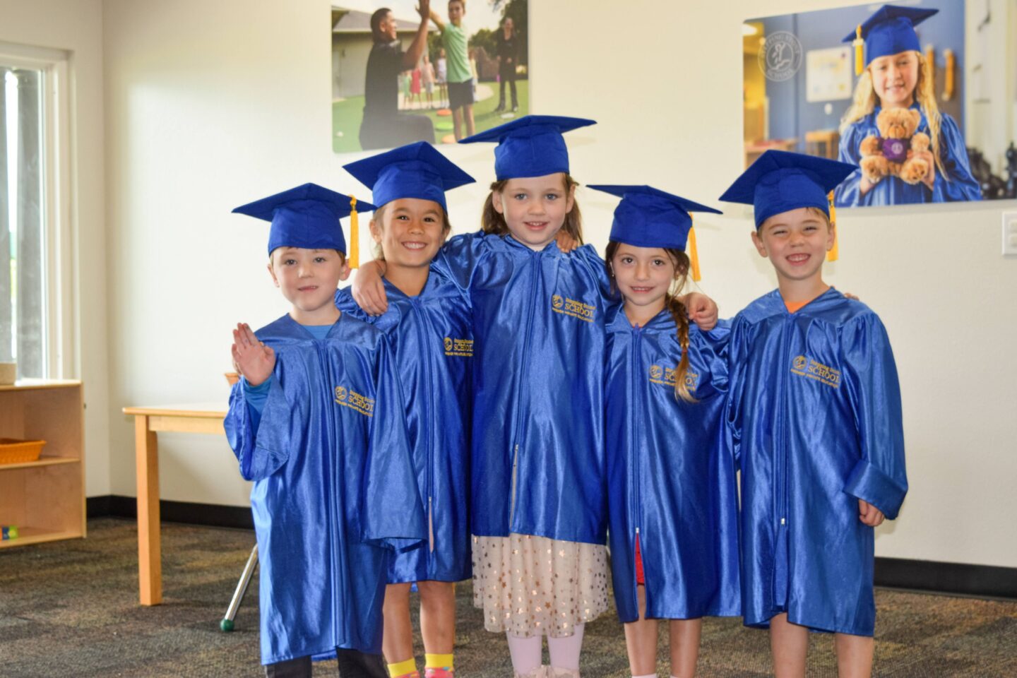 Five young graduates in blue caps and gowns celebrating in a classroom setting