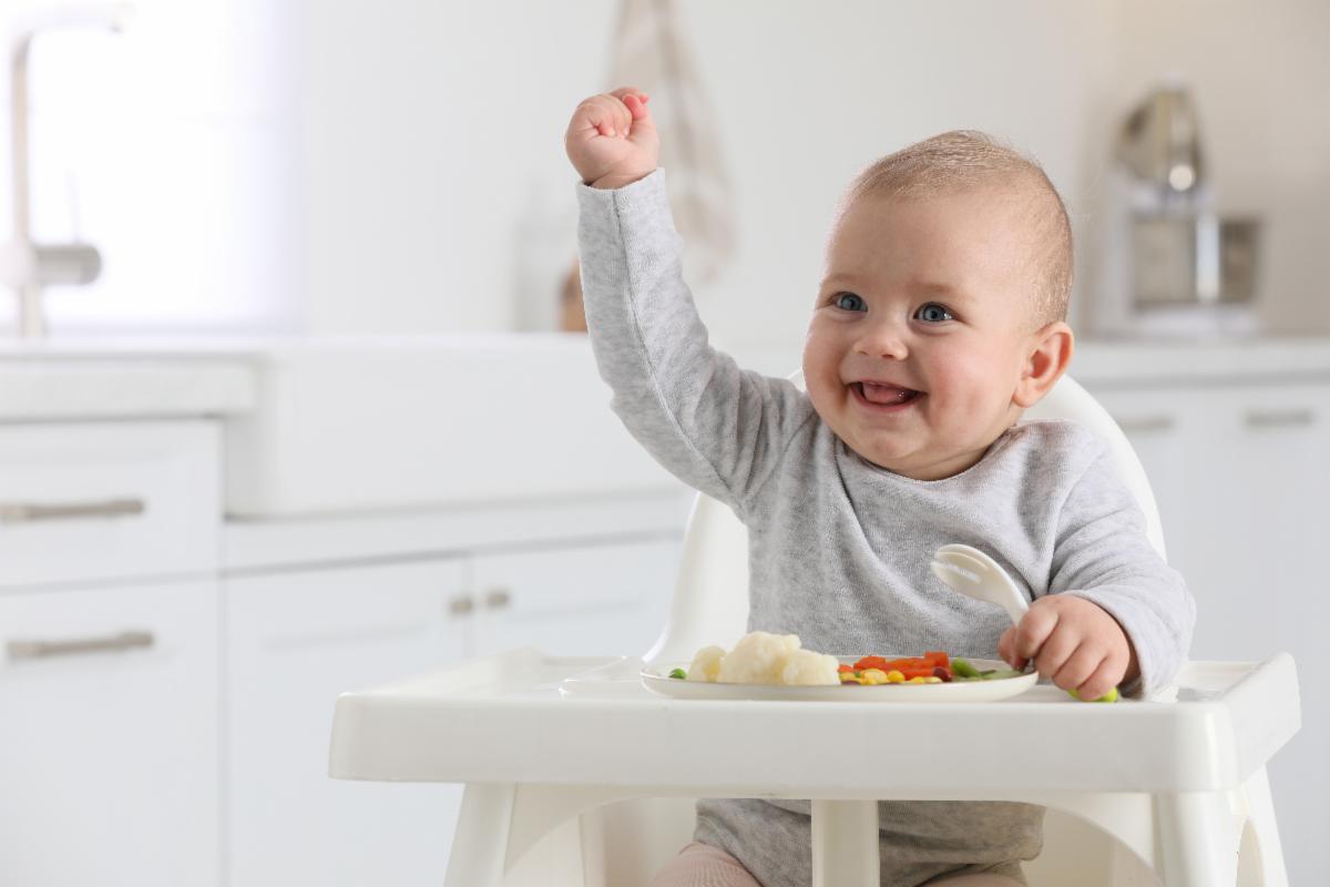 Smiling baby in a highchair with vegetables in a bright kitchen