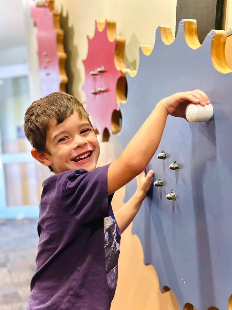 Young boy smiling while playing with interactive gear wall in a fun learning environment