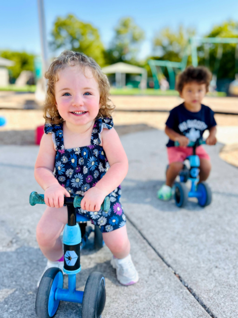 Smiling toddler riding a blue balance bike in a sunny playground