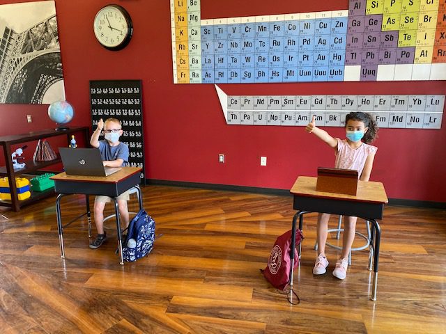 Children in masks sitting at desks in a classroom with periodic table on the wall.