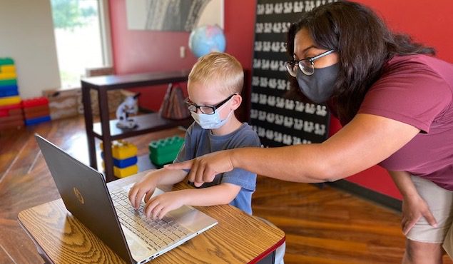 Teacher assisting a young student with a laptop in a colorful classroom both wearing masks