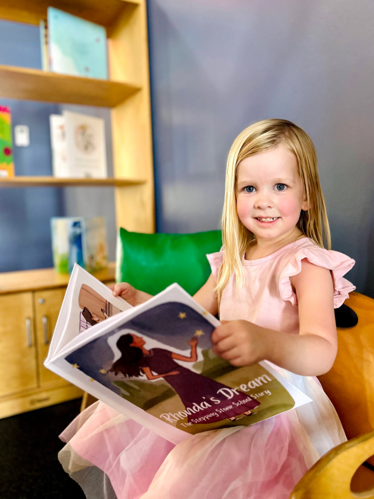 Smiling girl in pink dress reading a picture book in a cozy room with bookshelves