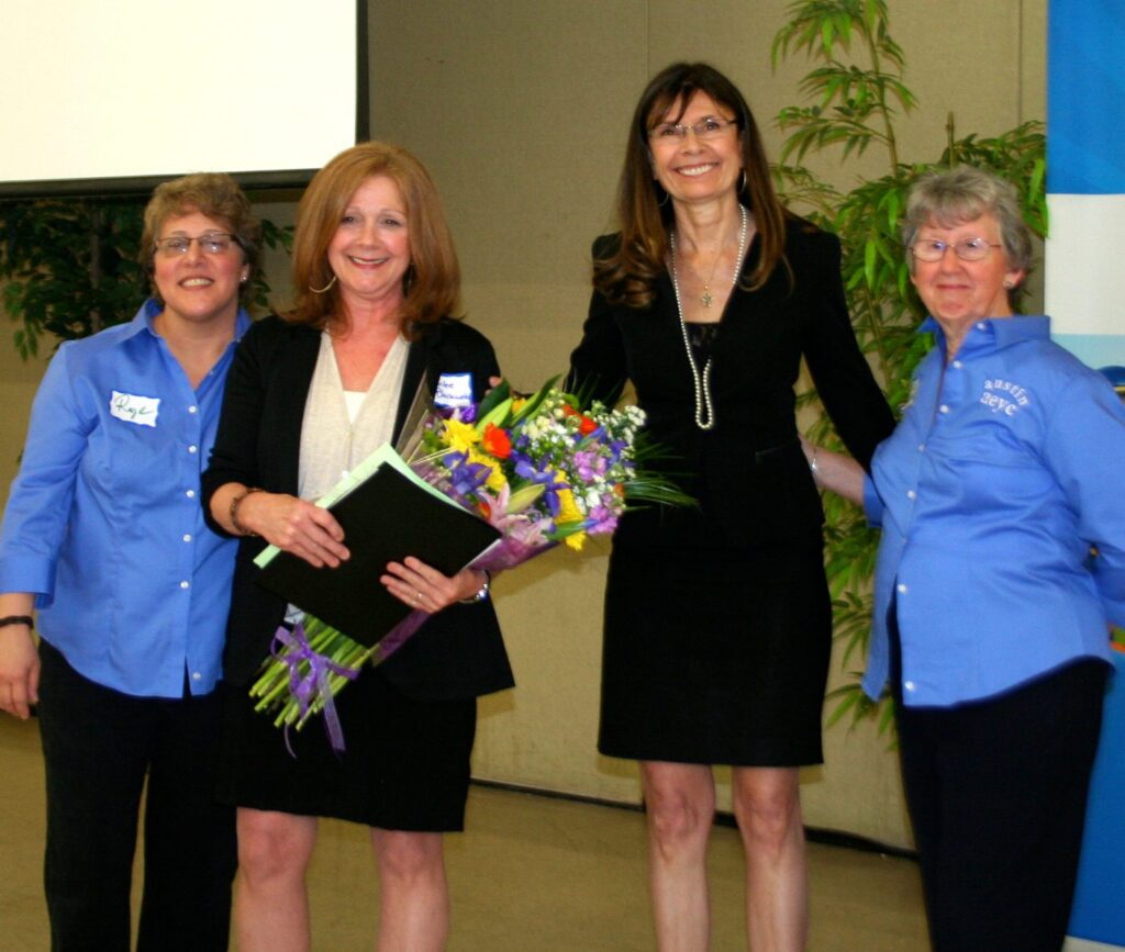 Four women smiling at an event one holding flowers indoors with a plant in the background