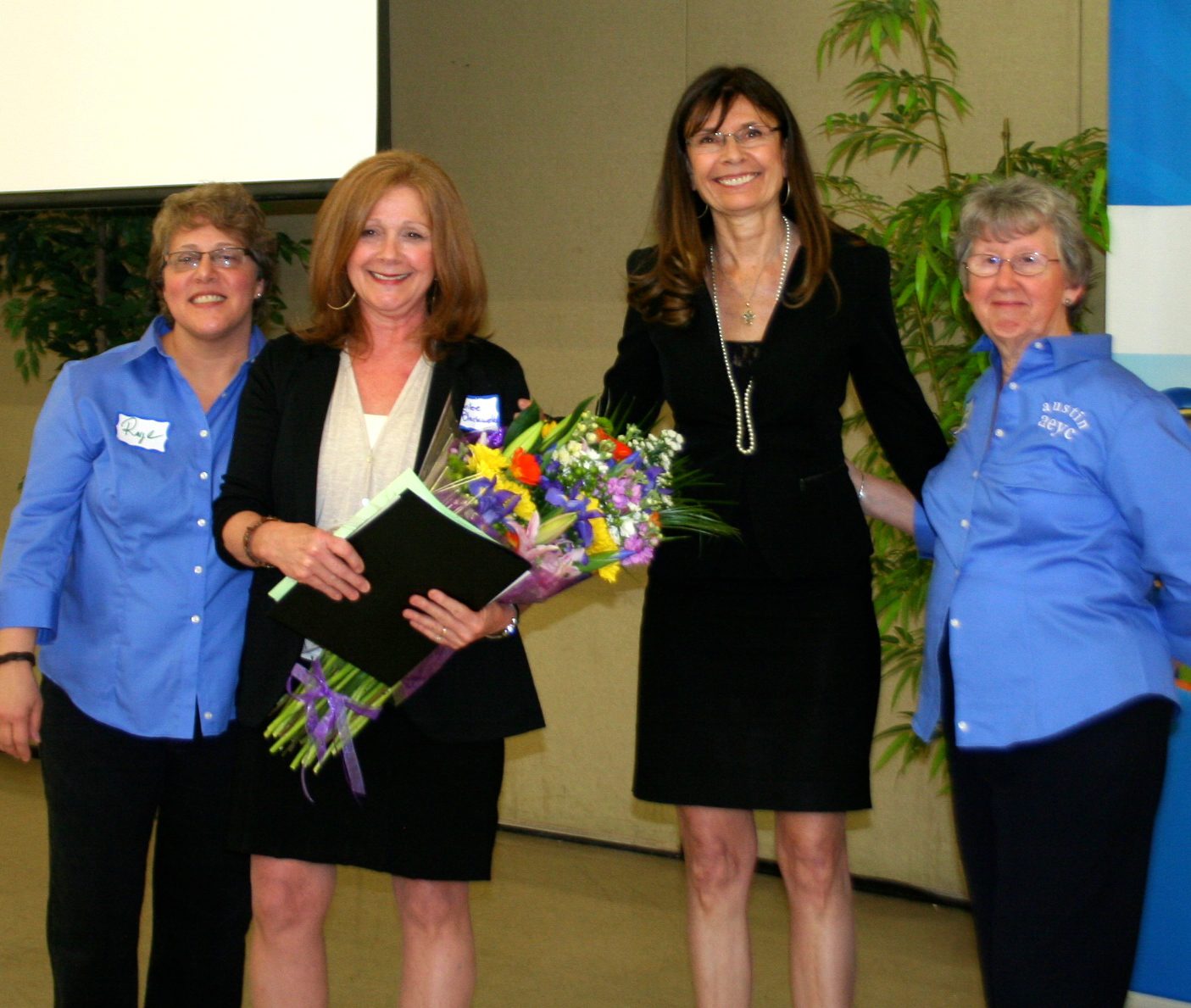 Four women smiling at an event one holding flowers indoors with a plant in the background