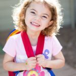 Smiling young girl with curly hair wearing a pink shirt and red backpack outdoors