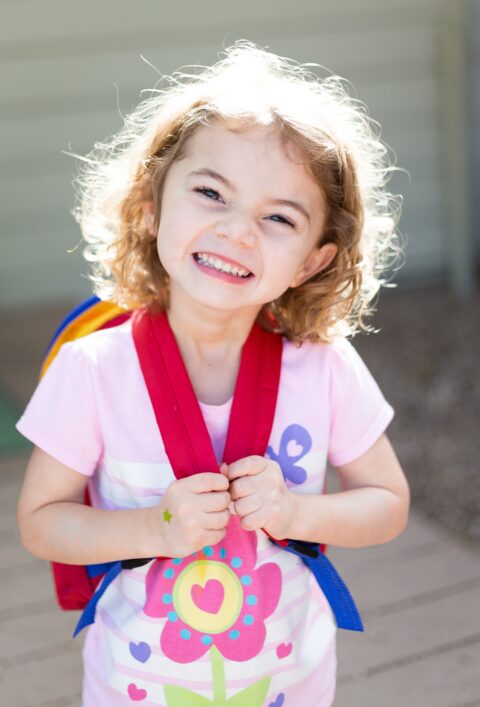 Smiling young girl with curly hair wearing a pink shirt and red backpack outdoors