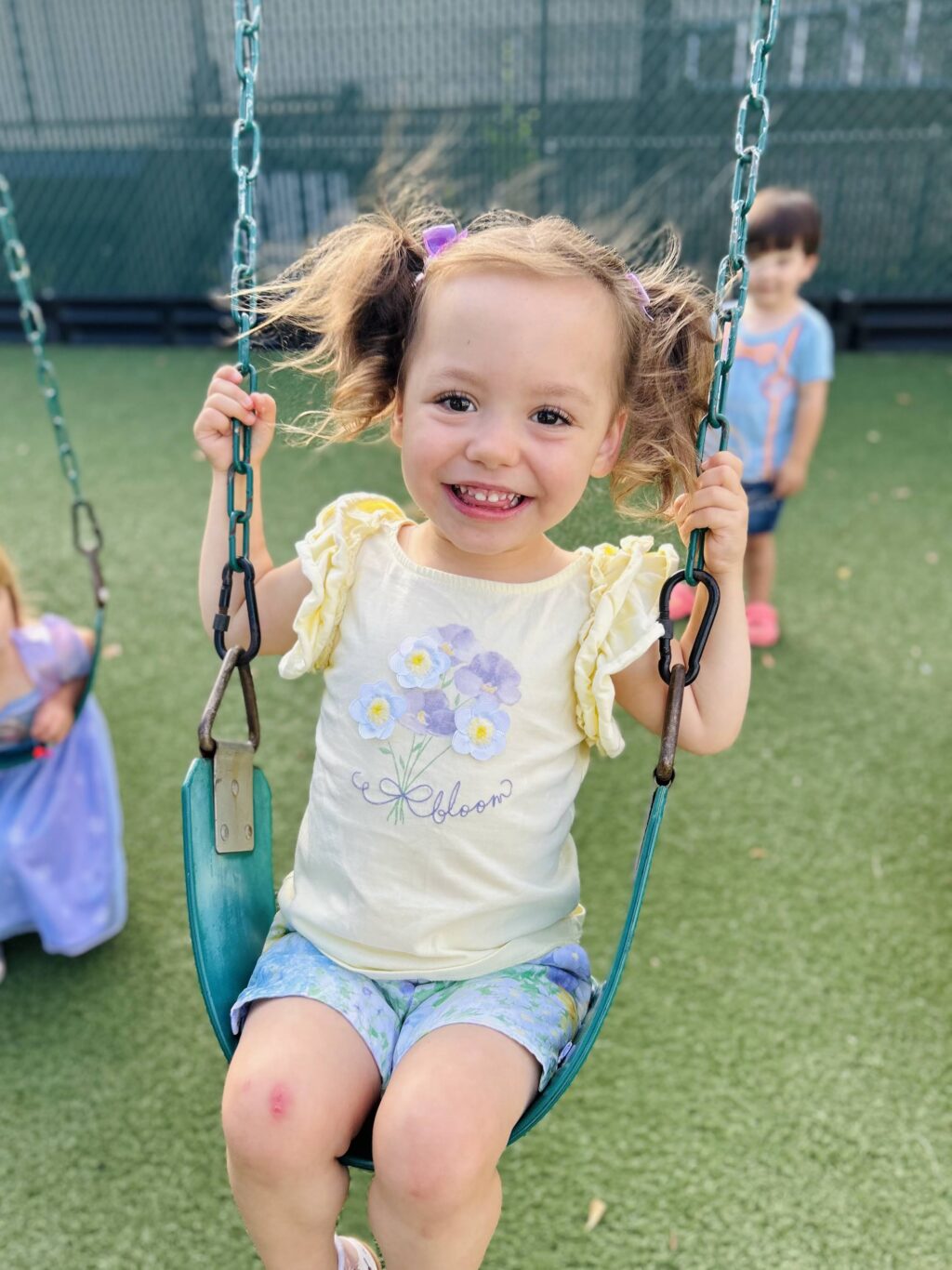 Smiling toddler on a swing wearing a floral shirt and shorts in a playground on a sunny day