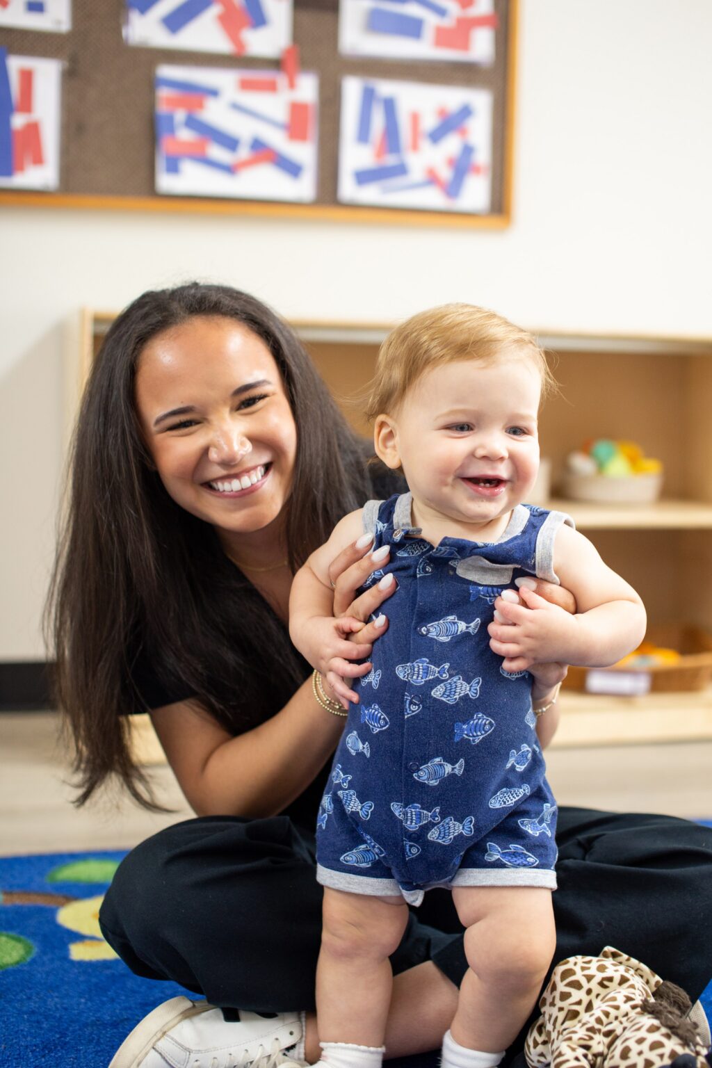 Smiling woman with a happy toddler in blue overalls in a classroom setting