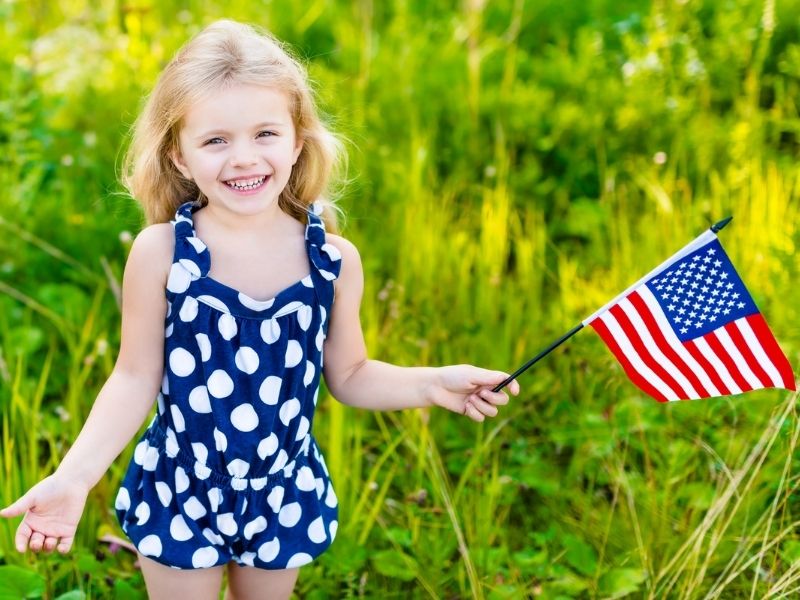 Smiling girl in blue polka dot dress holding American flag in a green field