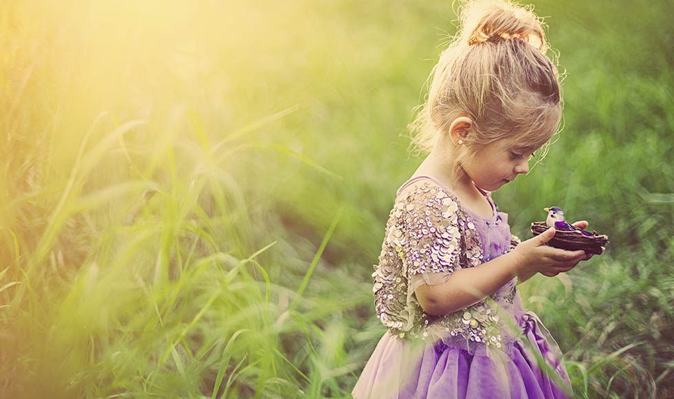 Young girl in purple dress holding a small bird's nest in a sunlit grassy field