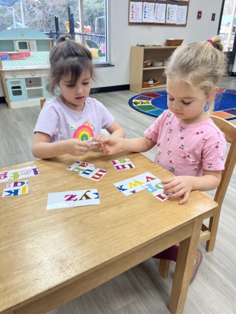 Two young girls at a wooden table playing with colorful alphabet cards in a classroom setting.