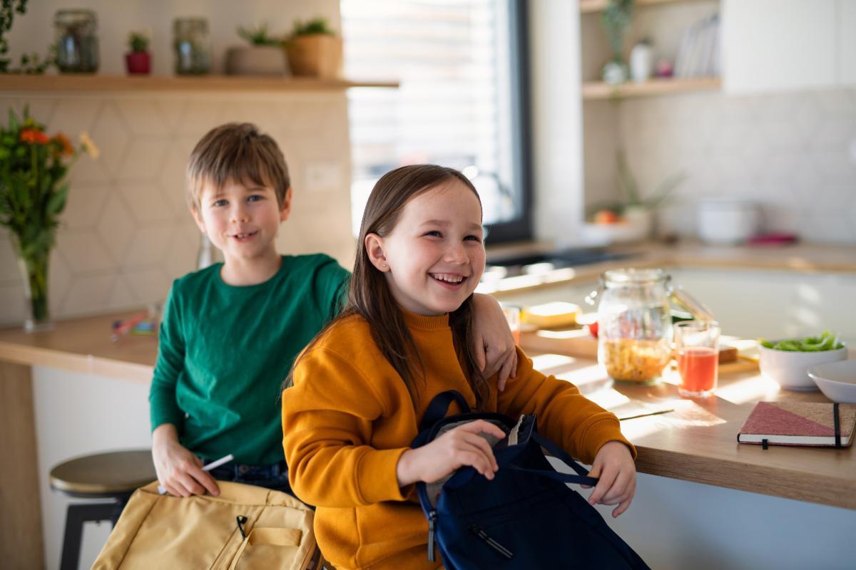Two smiling children with backpacks sitting in a bright kitchen ready for school