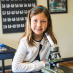 Young girl smiling next to a microscope in a classroom science setting.