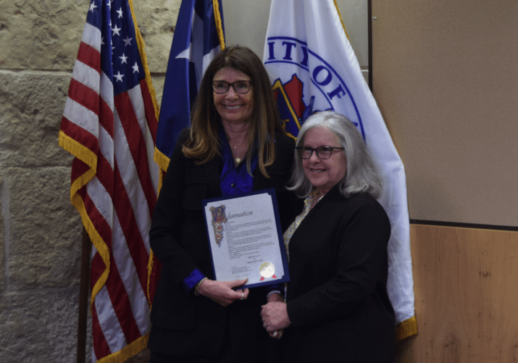 Two women hold a framed proclamation in front of American flags and a city emblem at an event.