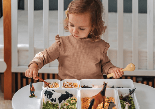 Toddler playing with sensory bins filled with grains and animal toys at a white table