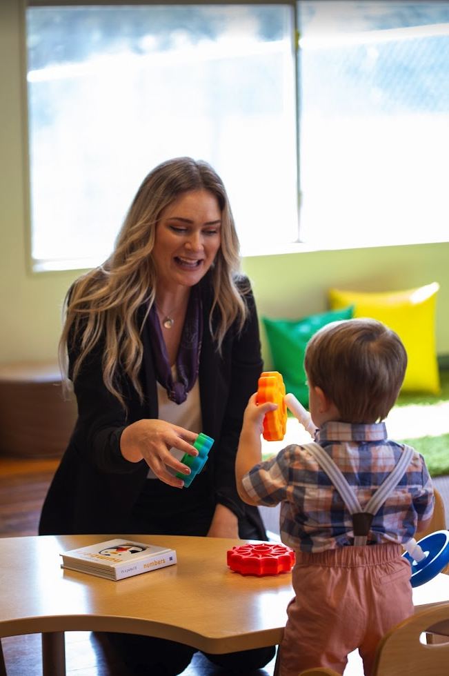 Teacher engaging with a young child using colorful building blocks at a table in a bright classroom.