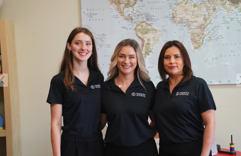 Three smiling women in black shirts stand in front of a world map in an office setting.