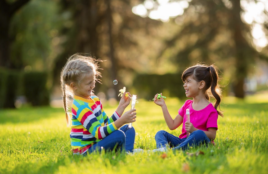 Children blowing bubbles and laughing while sitting on grass in a sunny park.
