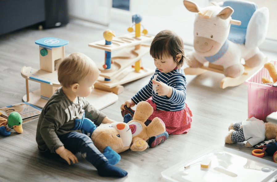 Toddlers playing with toys on a soft rug in a bright playroom with stuffed animals and wooden toys nearby