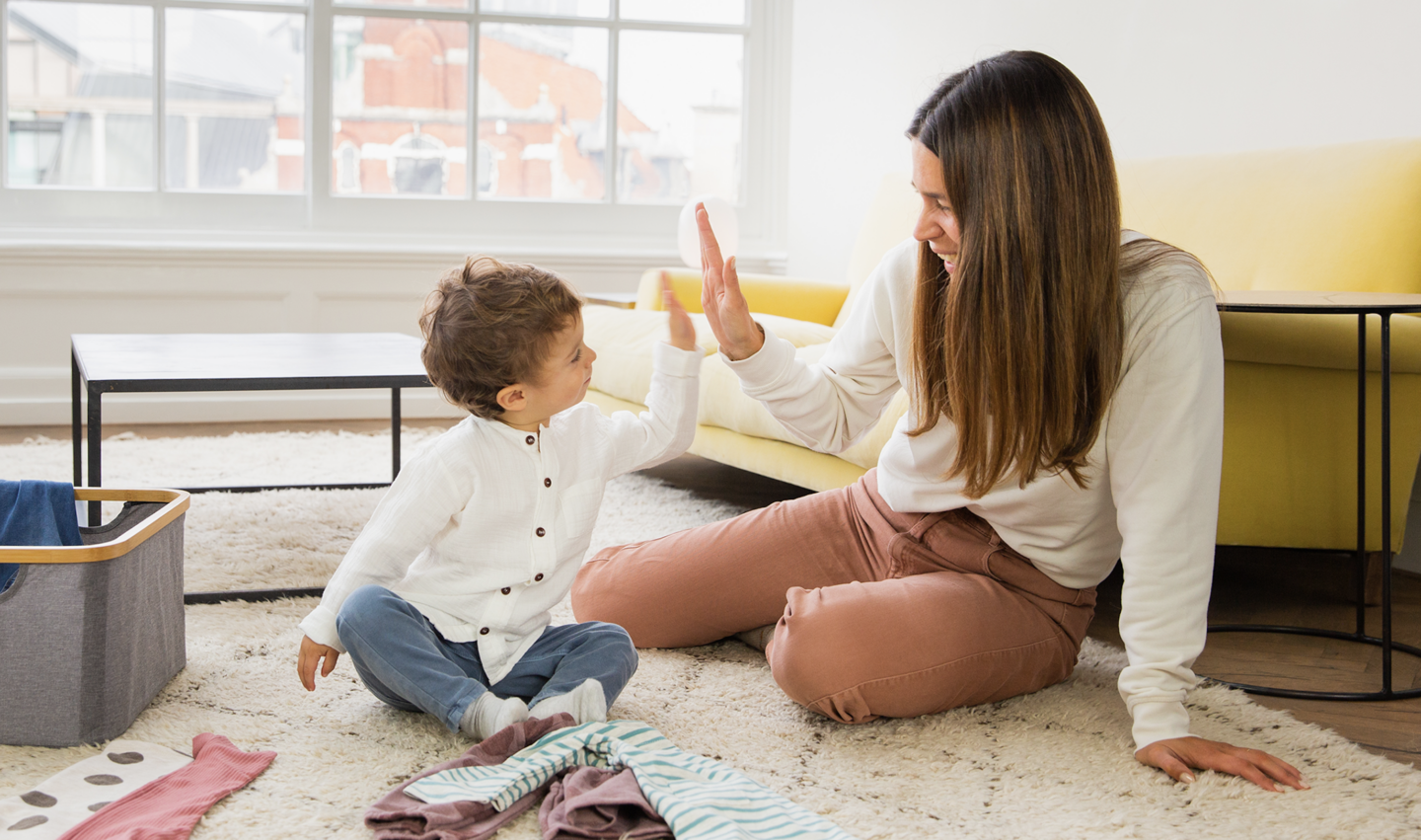 Mother and child high-fiving on living room floor with laundry nearby