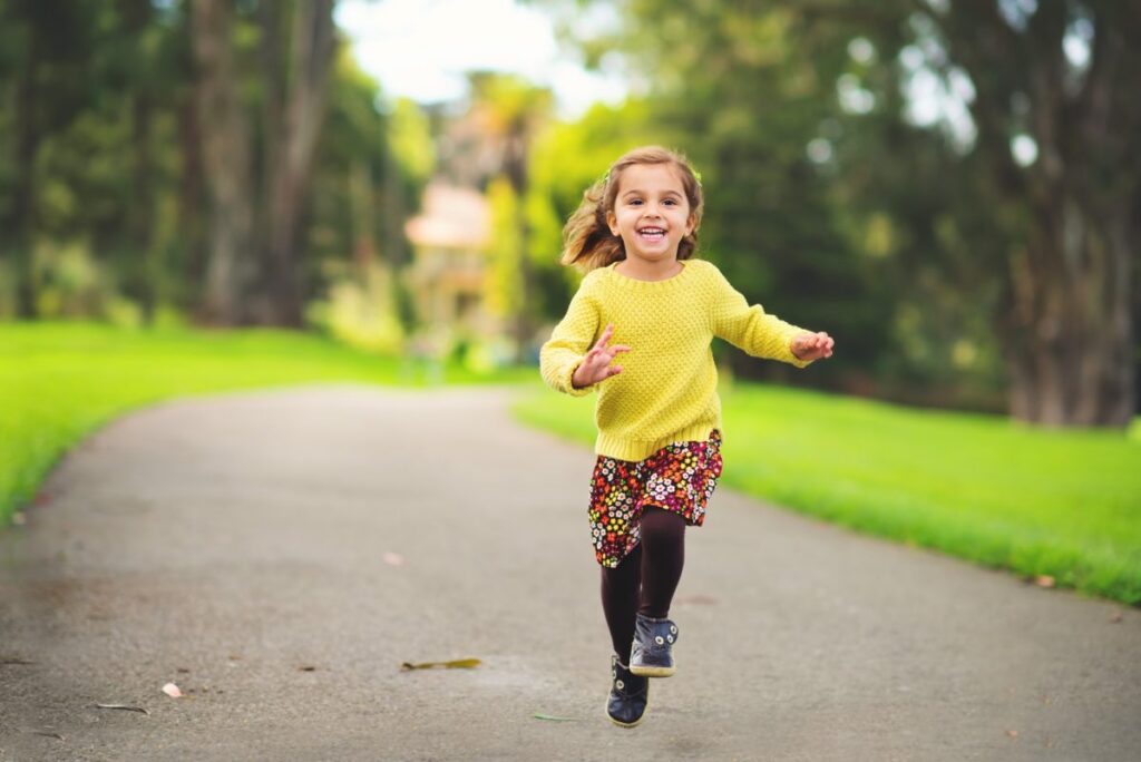 Young girl in yellow sweater joyfully running on a park path.