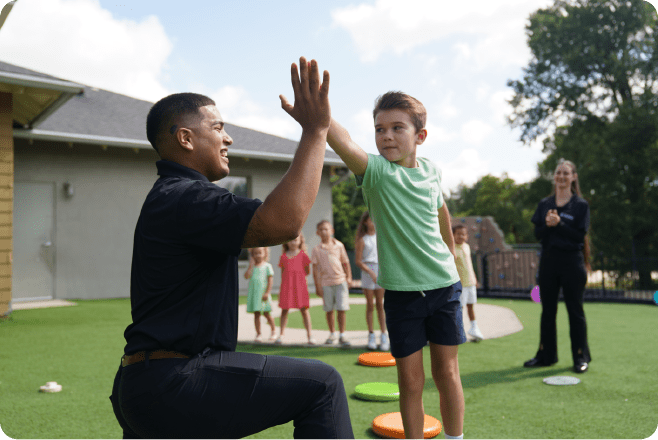 Child high-fives teacher outdoors on playground with other children in the background