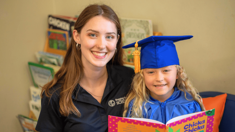 Smiling woman with young girl in graduation cap reading a book together in a classroom setting