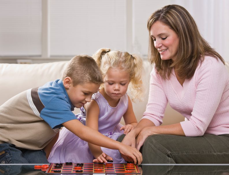 Mother playing checkers with her two children on a cozy living room sofa.