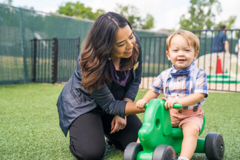 Woman and toddler smiling outdoors with a green toy vehicle in a playground setting