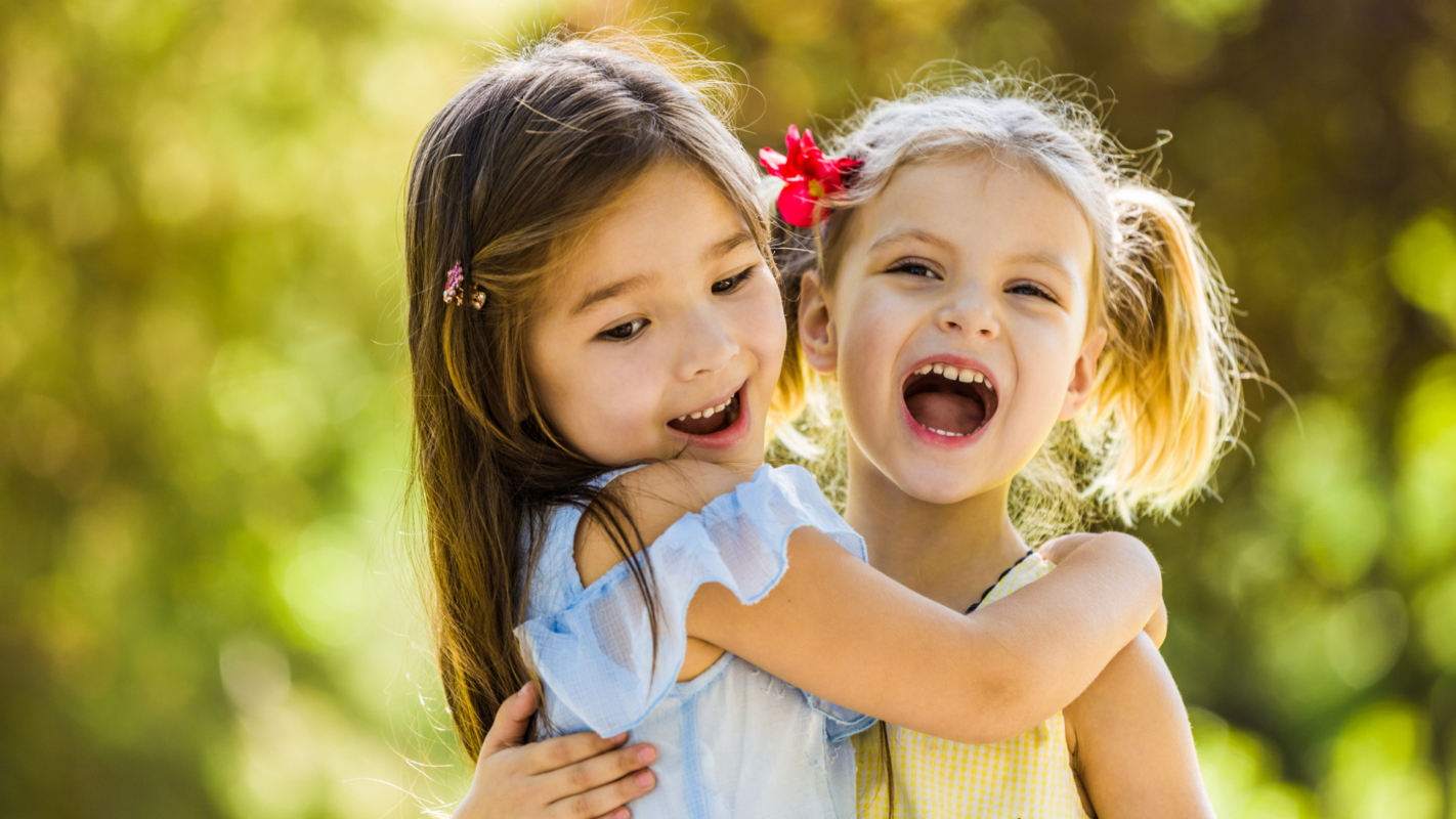 Two young girls hugging and laughing outdoors in a sunny park