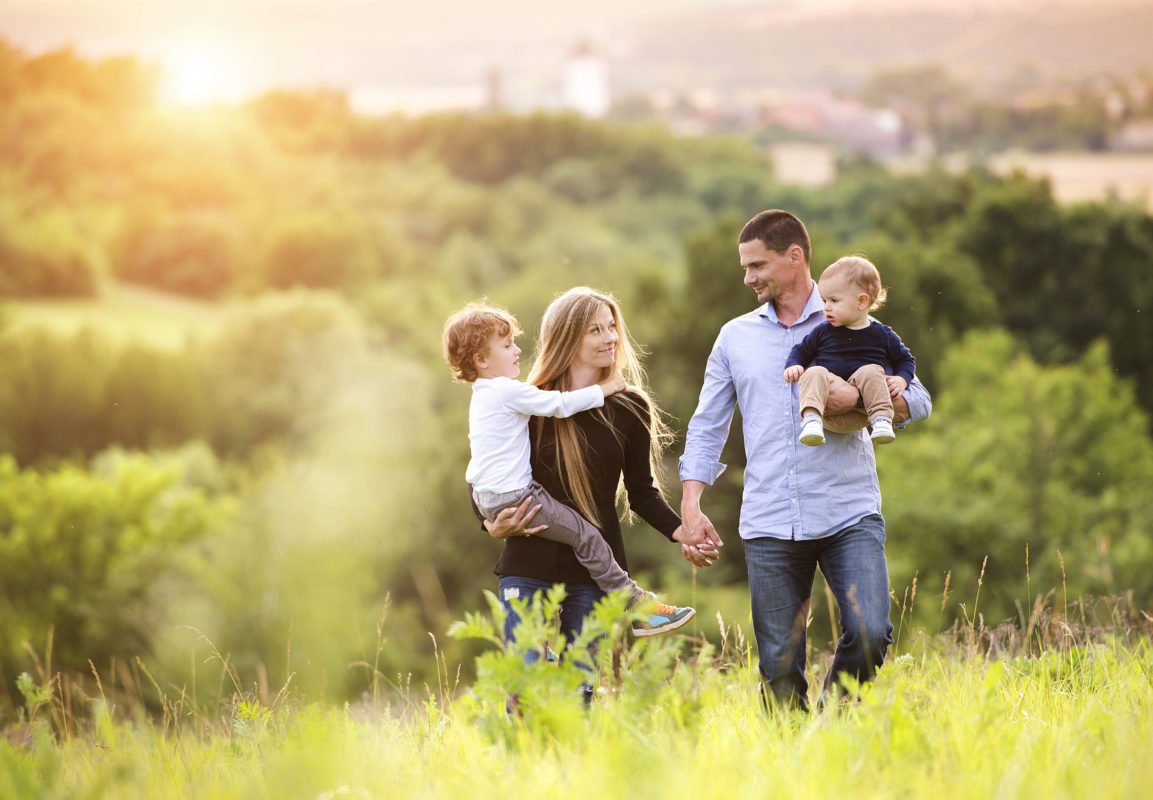 Family walking in sunny field holding children surrounded by lush greenery