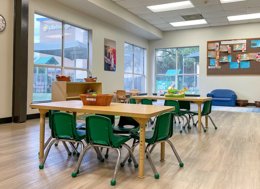 Bright classroom with tables, green chairs, and educational decor, featuring large windows and wooden floor.