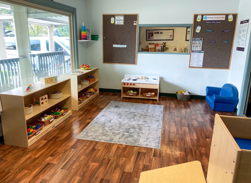 Bright classroom for young children with shelves, learning toys, and a play area on a wooden floor.