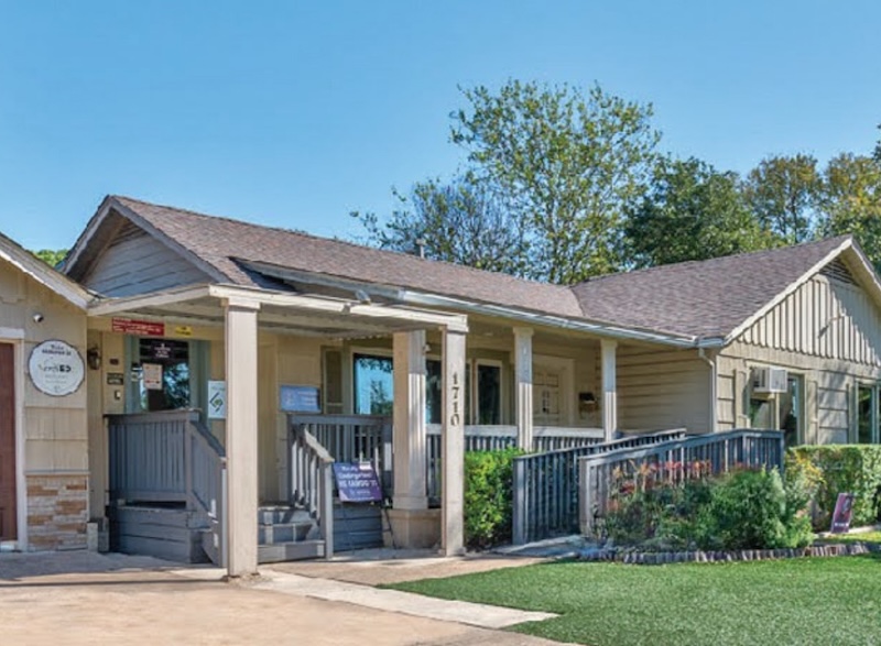 Small office building with a sloped roof and front porch surrounded by greenery on a sunny day
