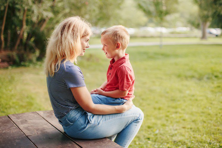 Mother and young son smiling at each other while sitting on a picnic table in a green park